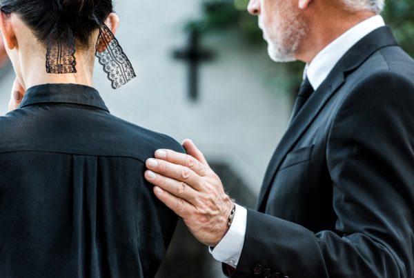 older man in funeral attire placing hand on shoulder of young woman in black blouse with black lace ribbon in hair