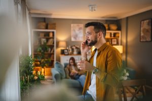 Man talking on phone to a grieving friend; wife sitting on couch in background
