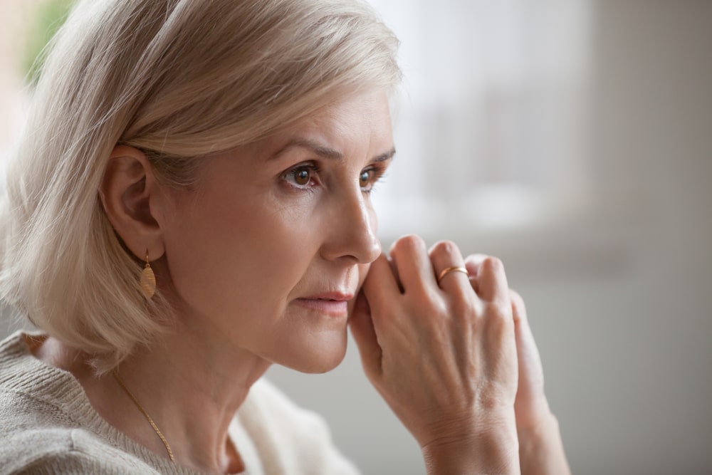 older woman staring thoughtfully while clasping her hands by her face.