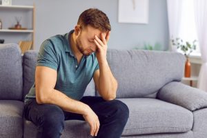 Young man sitting on couch at home with hand on his face; experiencing grief & depression