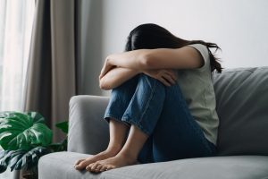 Young woman sitting on couch with knees pulled to chest and arms crossed over knees; grief & depression