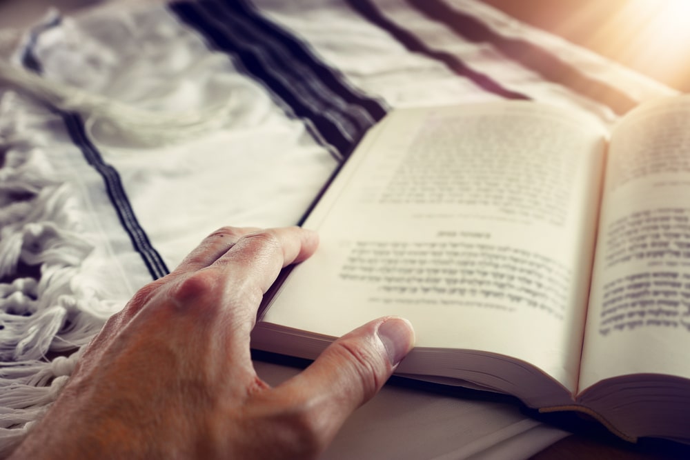 Man's hand touching a prayer book resting on top of a Jewish prayer shawl