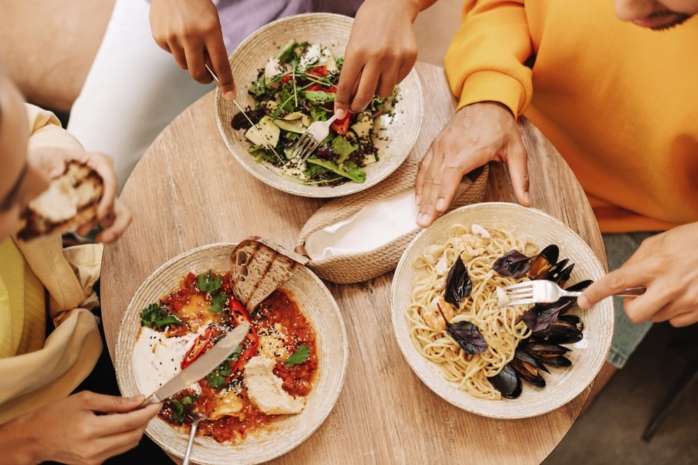 Three people eating out at a restaurant with three different pasta dishes in front of them on a round wooden table