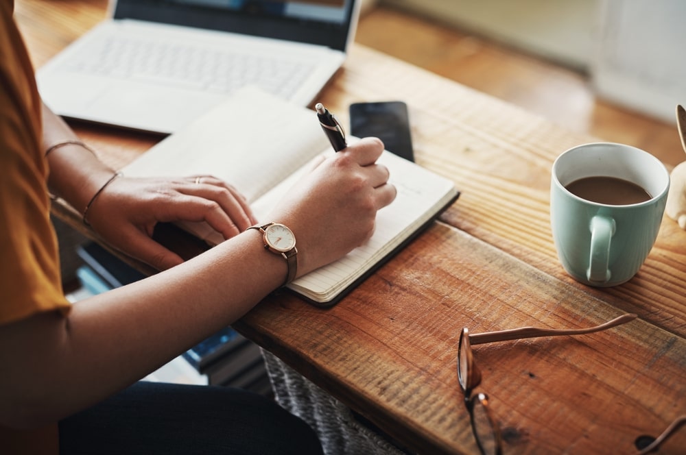 woman writing a eulogy in a notebook on a wooden table next to a laptop and a cup of coffee