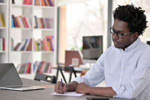 man with glasses writing on a piece of paper with a laptop next to him on the table