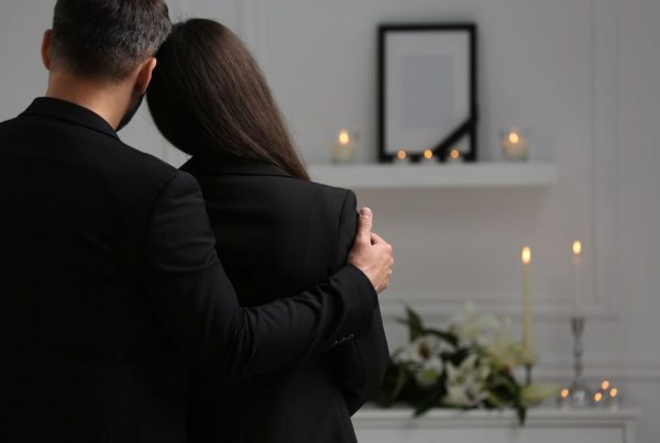 Man and woman in black clothing leaning on each other in a comforting way as they attend a visitation