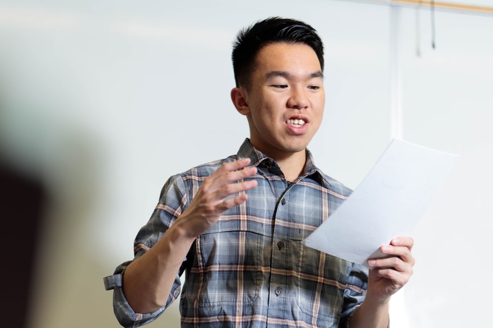 Young man in a plaid shirt practicing a eulogy by reading it aloud from a paper
