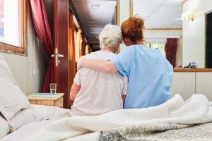Mature woman and her nurse sitting on the edge of patient's bed, sharing a comforting side hug
