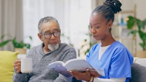Older man and hospice nurse sitting on couch in his home with educational book