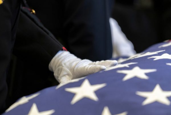 Uniformed military personnel resting hand on flag-draped casket