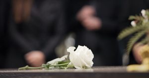 a white rose on top of a closed casket with people blurred in the background