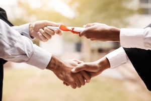 Two people's hands, doing a handshake and passing off a diploma