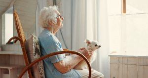 elderly woman holding a small dog while looking out a window