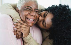 Woman with arms around her mother's neck, kissing her on the cheek.