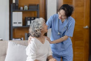 Healthcare worker standing beside a woman on a couch, with her hand on her shoulder.