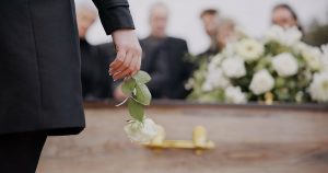 Mourner holding a white flower as they look at a wooden casket with a spray of white flowers on top