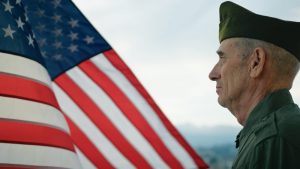 Older man in uniform standing and facing an American flag