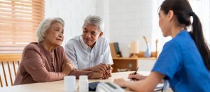 An elderly couple and a healthcare worker sitting at a table and talking.