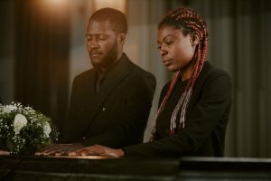 Grieving man and woman standing next to a casket, hands rested on its lid