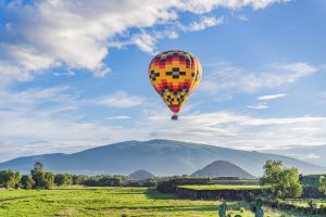 Multi-colored hot air balloon hovering over a field with mountains behind