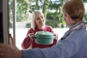 A woman holding a covered dish while another woman opens her front door.