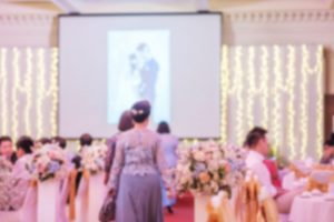 A wedding reception with people sitting at tables or walking around, a photo on display on a screen in the background