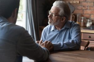 Two men sitting at a table holding hands while one gazes out the window.