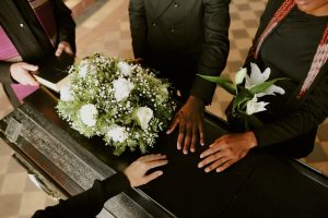 Close up of hands placed on a casket beside a bouquet of white flowers, and someone holding a bible off to the side