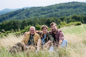 grandad, grandson, and son sitting in a field together with hills behind them