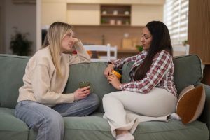 Two women sitting on a couch, holding coffee mugs and talking.