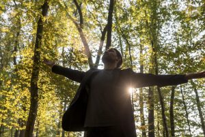Man with arms outstretched in the middle of the woods, looking up at the sky.