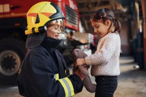 Firefighter crouched down talking to a little girl holding a stuffed animal.