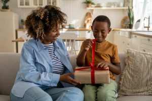 Woman and child sitting on a couch together while he unwraps a gift in his lap.