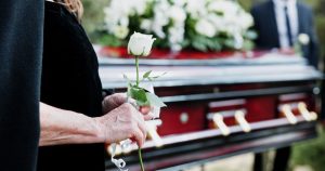 Woman in black dress holding white rose while standing next to wooden casket
