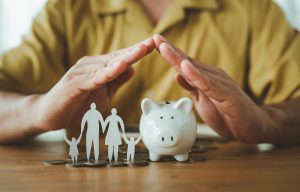 Man in a yellow shirt creating a roof with his hands over a paper cutout of a family and a ceramic piggy bank