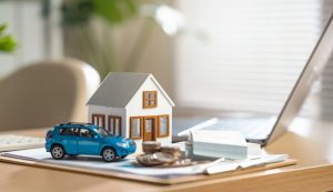 Toy house and toy car sitting on a desk next to a laptop and stack of coins