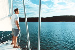 Man in shorts and polo standing on bow of a sailboat, looking out at the water